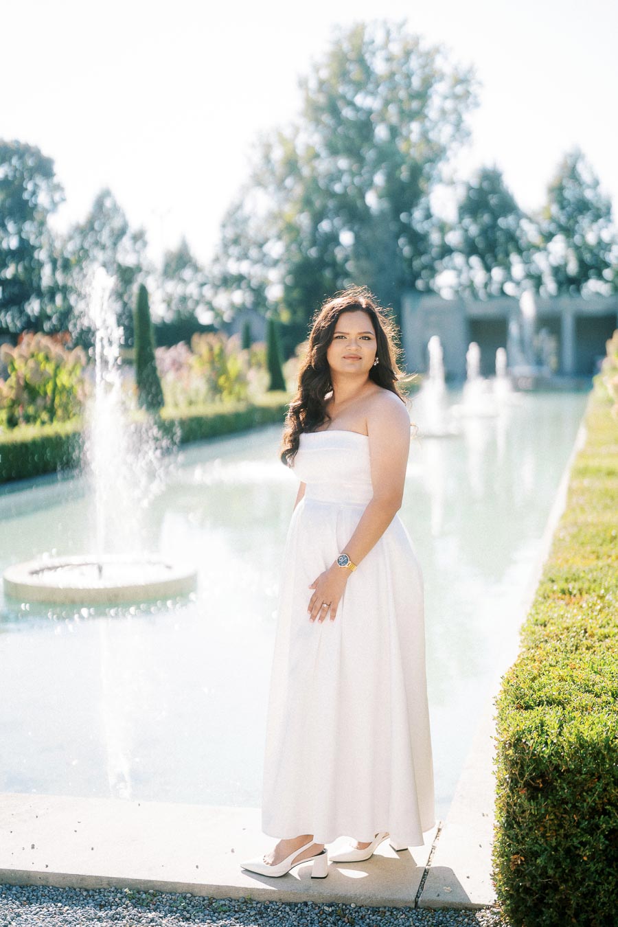 A woman in a flowing white dress stands by a serene fountain in a beautifully landscaped garden under a clear blue sky.