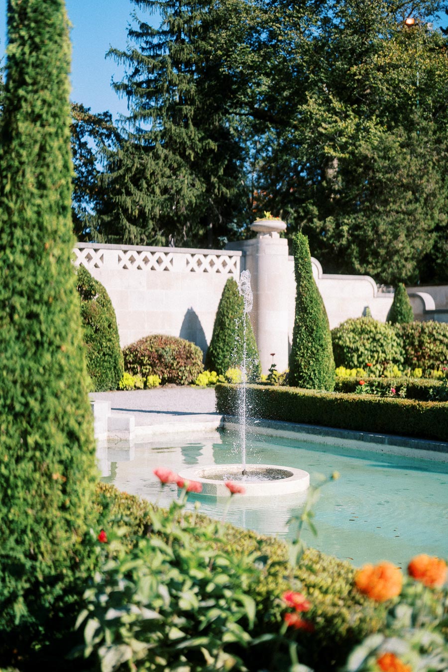 Elegant garden with a central fountain surrounded by neatly trimmed hedges and vibrant flowers, set against a backdrop of lush trees and blue sky.