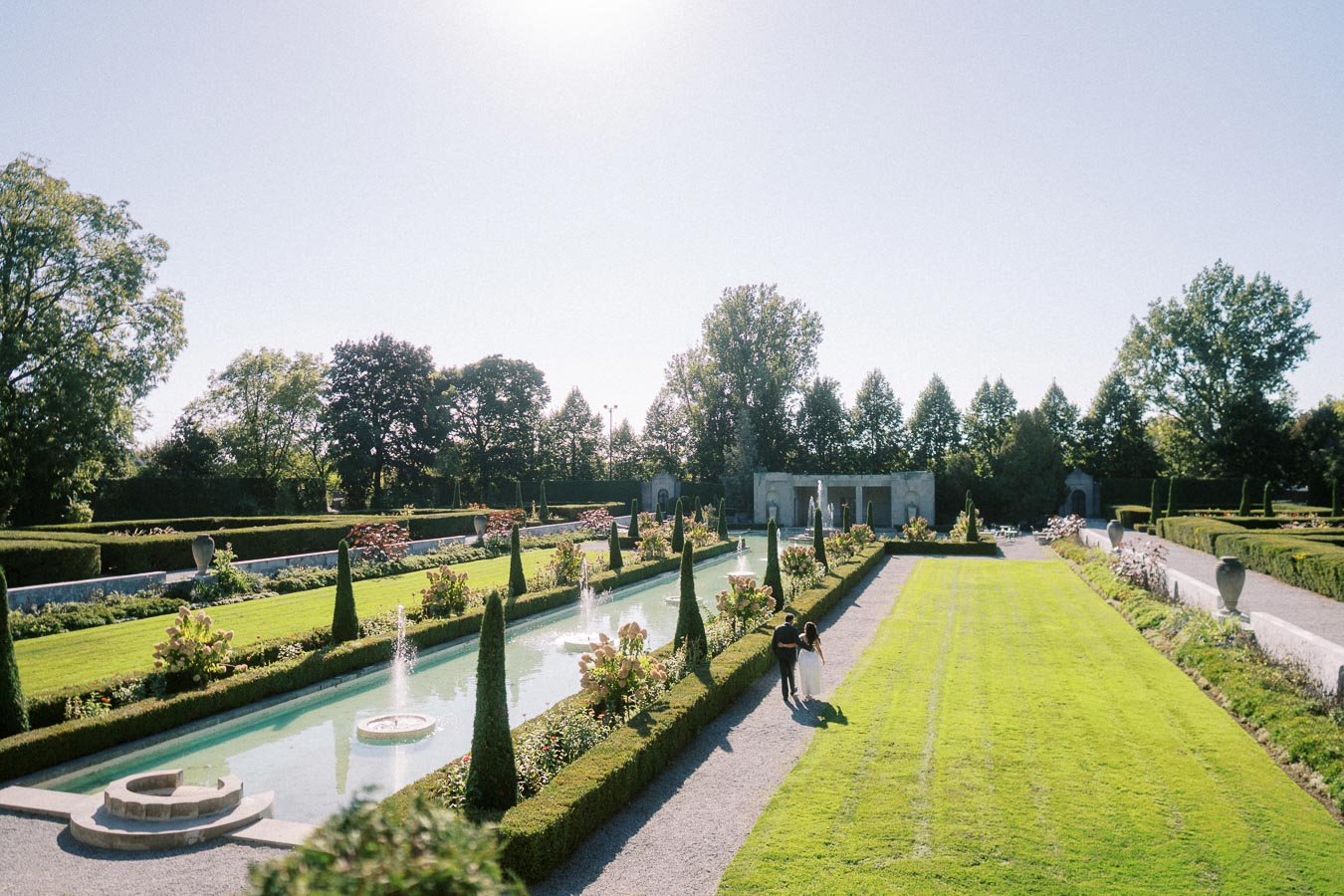 Luxurious garden with symmetrical hedges and fountains under a clear blue sky, featuring a couple walking along a gravel path, surrounded by vibrant greenery and well-maintained landscaping.