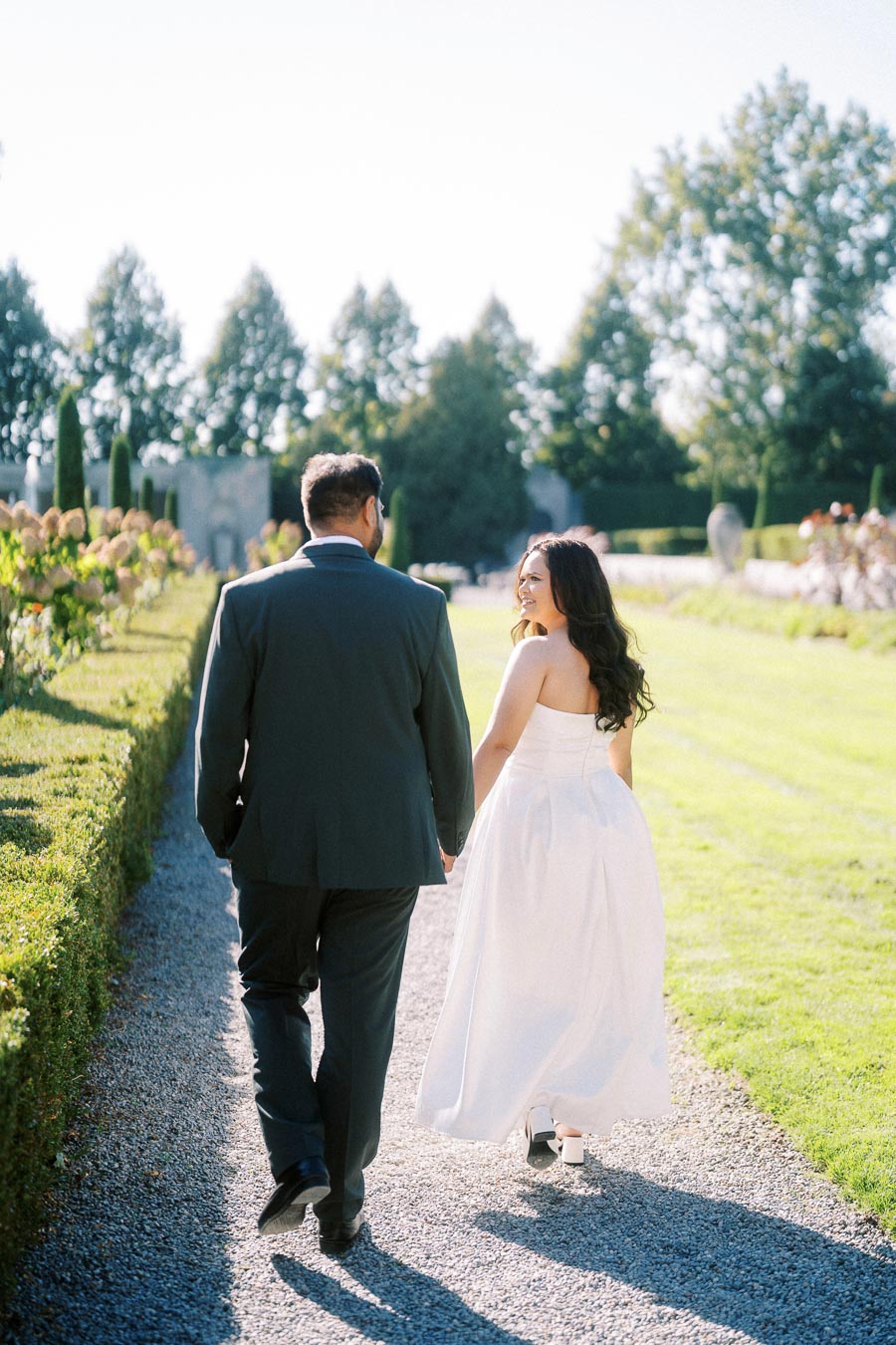 Elegant couple walking on a garden path during a sunny day in formal wedding attire, surrounded by lush greenery and well-manicured hedges.
