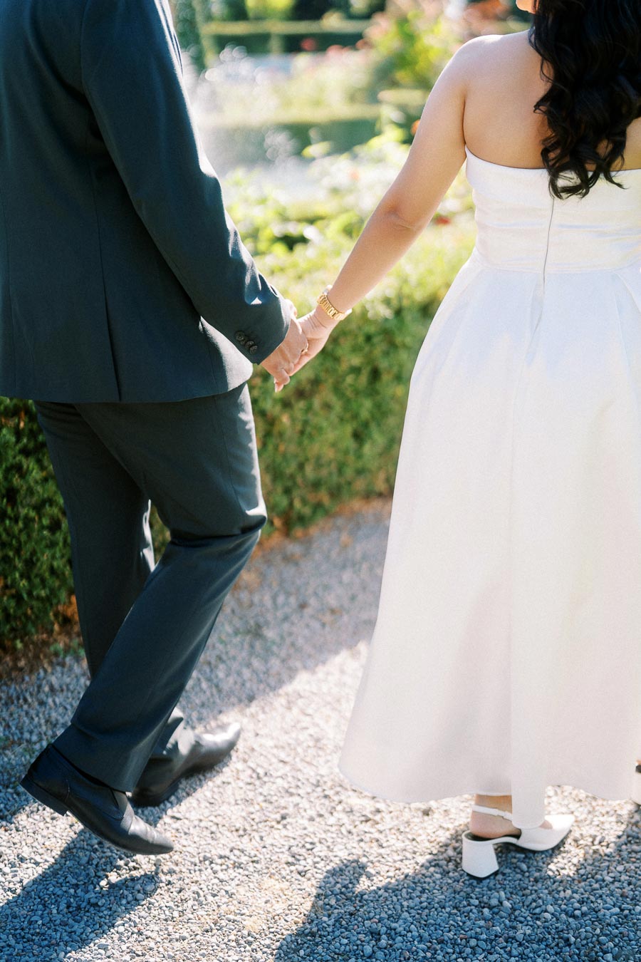 A couple holding hands in formal attire, walking on a gravel path in a sunlit garden. The woman is wearing a white dress and the man is in a dark suit.