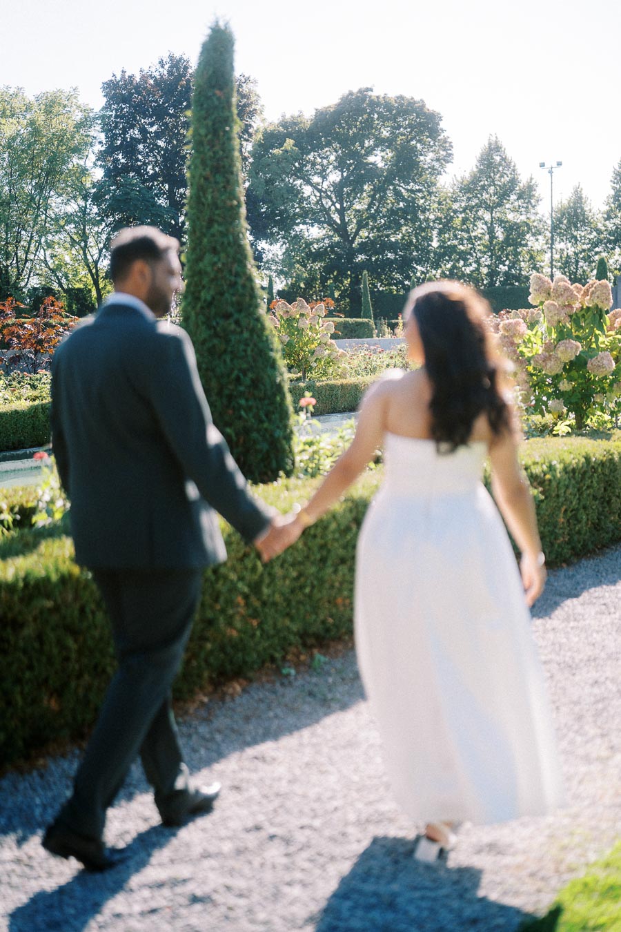 A couple holding hands and walking through a sunlit garden, with well-trimmed hedges and colorful flowers surrounding them. The man is in a dark suit, and the woman is wearing a white dress.