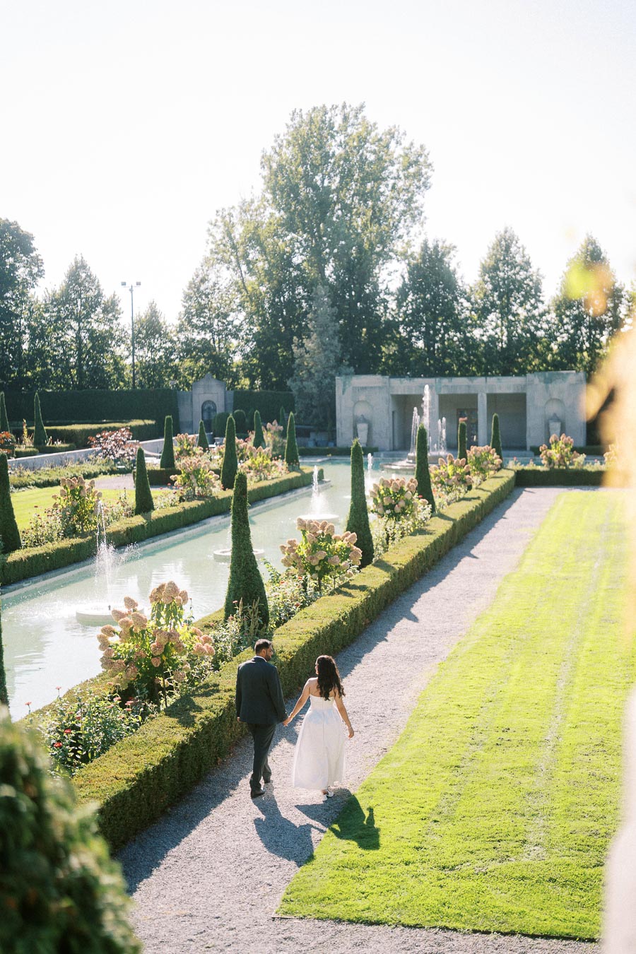 A couple holding hands walks along a picturesque garden path lined with fountains and manicured bushes, surrounded by vibrant greenery on a sunny day.