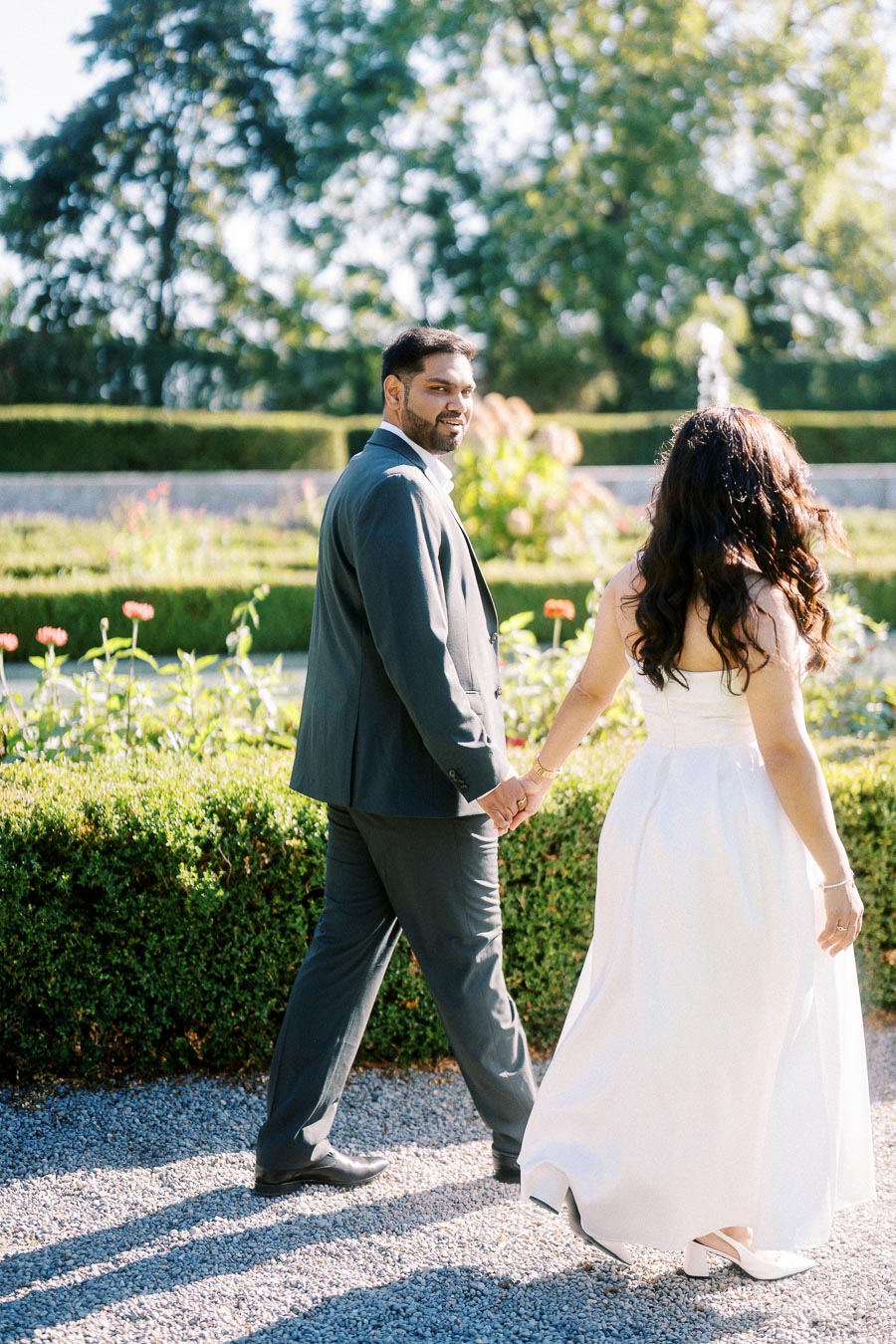 A couple walking hand in hand through a sunlit garden, with the man in a suit and the woman in a white dress, capturing a romantic moment surrounded by greenery and flowers.