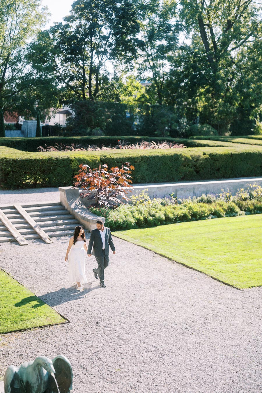 A couple in formal attire walking hand in hand through a beautiful garden with neatly trimmed hedges and vibrant greenery on a sunny day.