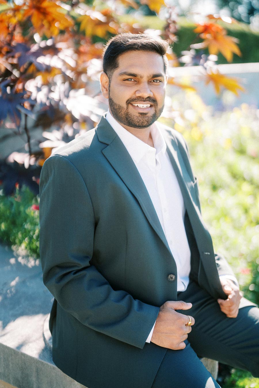 Man in a suit smiling outdoors, with autumn leaves and greenery in the background.