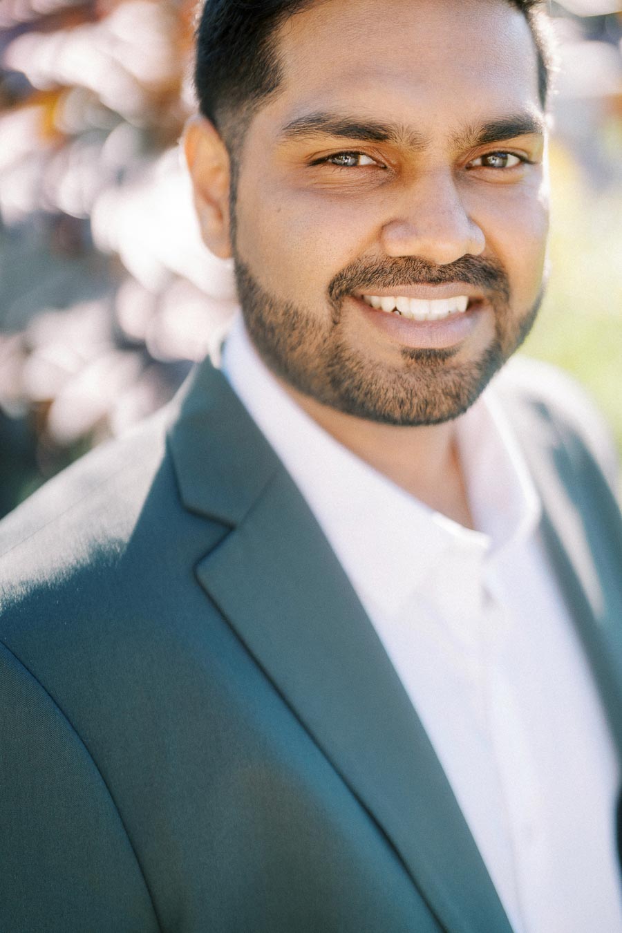 A smiling man wearing a green suit jacket and white shirt, standing outdoors with a blurred natural background.