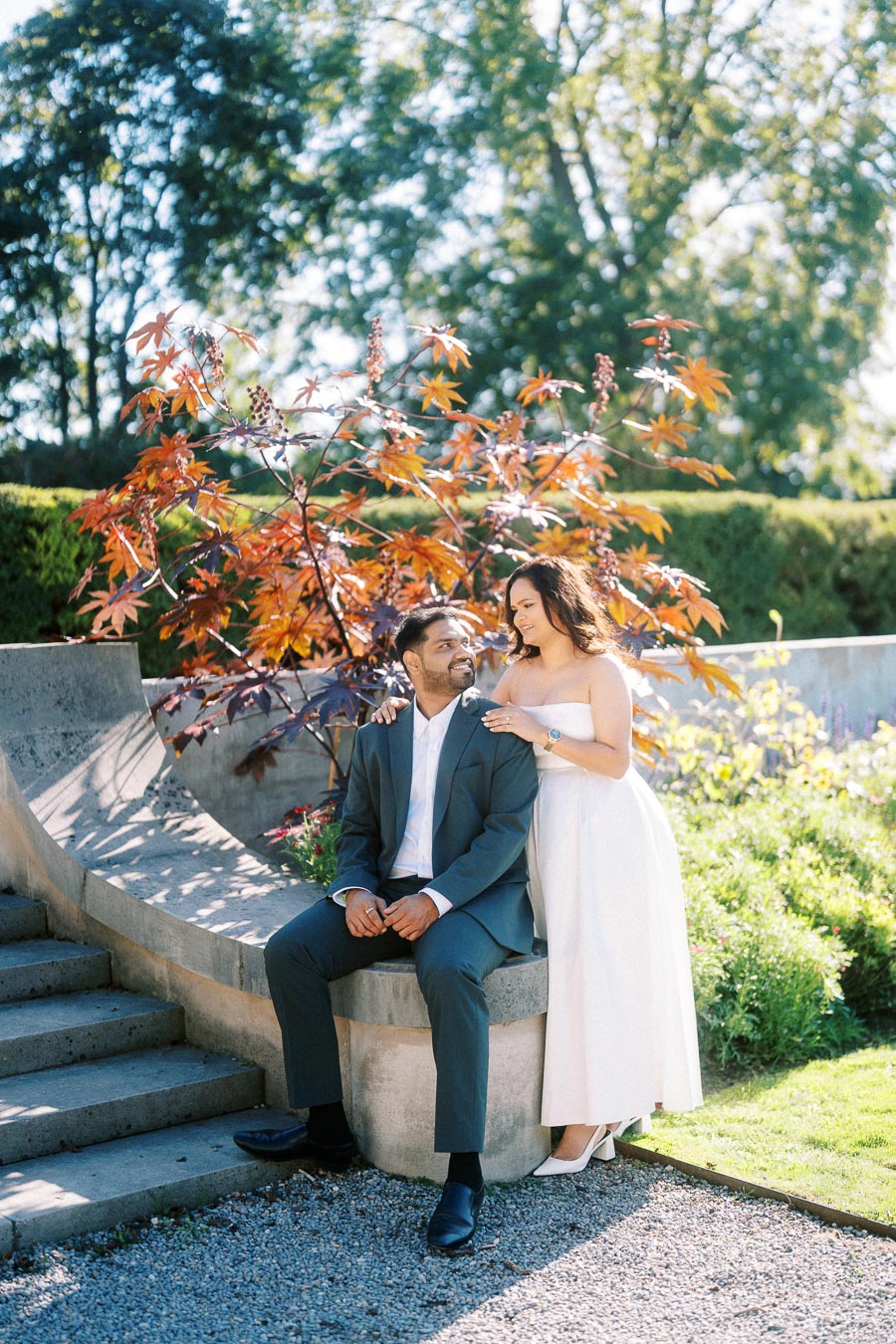 A couple posing in a sunny garden setting with vibrant autumn foliage and stone steps. The man is seated in a suit while the woman, in a white dress, leans affectionately over him.