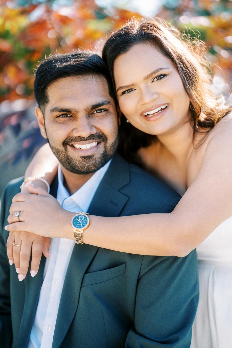 A happy couple smiling in an outdoor setting with fall foliage; the woman embracing the man from behind, both dressed elegantly.