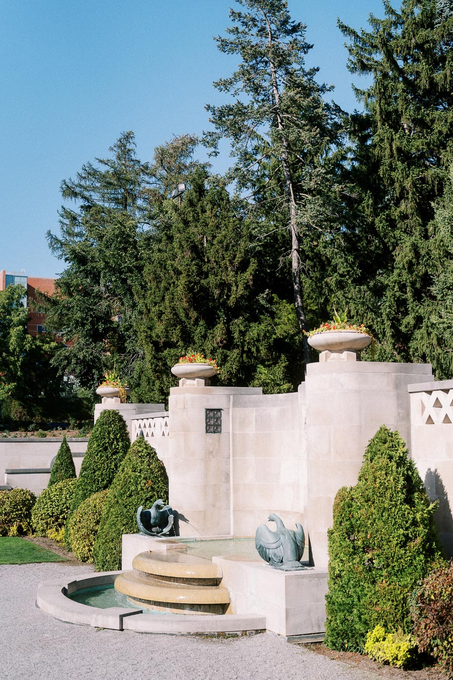 Serene garden view featuring a stone wall adorned with large planters filled with vibrant flowers, surrounded by lush greenery and tall trees under a clear blue sky. In the foreground, a decorative fountain enhances the tranquil ambiance of the landscape.