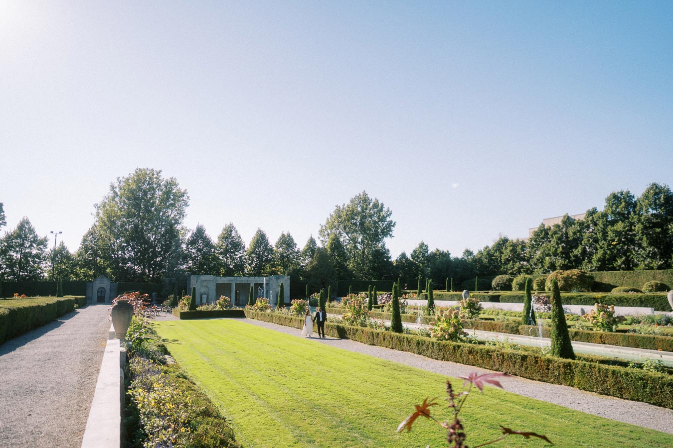 Elegant garden landscape with trimmed hedges and vibrant flowerbeds under a clear blue sky, featuring a couple walking along a gravel path amidst lush greenery.