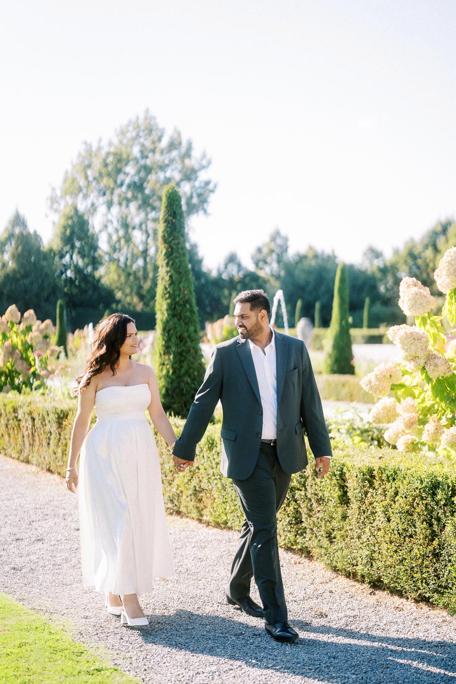 Romantic couple walking hand in hand through lush gardens on a sunny day, both elegantly dressed, with vibrant greenery and flowers in the background.
