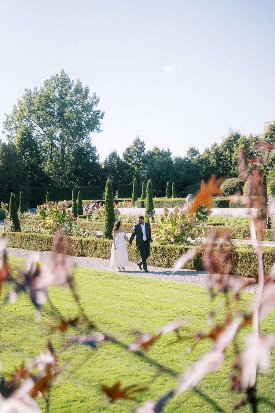 A bride and groom walking hand in hand through a picturesque garden on a sunny day, surrounded by lush greenery and colorful flowers, symbolizing romance and tranquility.