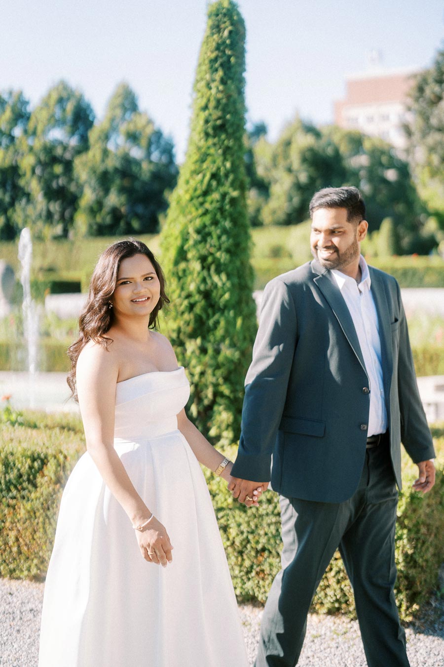 A couple holds hands and walks together in a lush garden, with the woman wearing a white dress and the man in a dark suit.