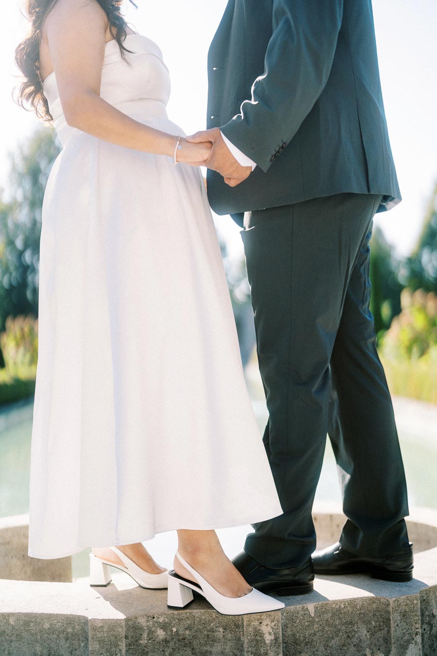 Elegant wedding couple holding hands in sunshine, bride in white dress and groom in dark suit, outdoor setting