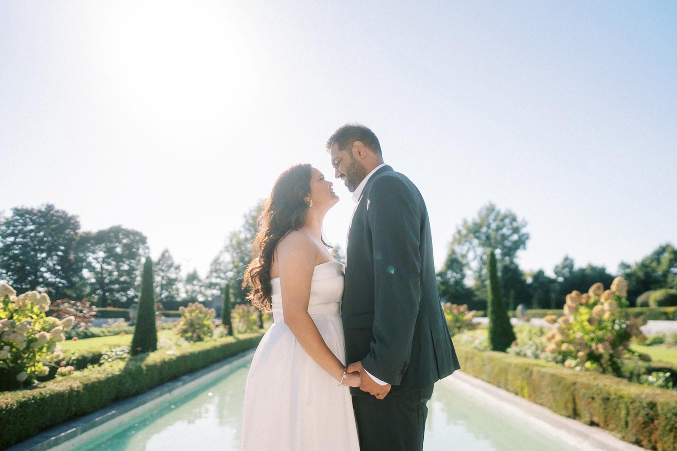 Romantic wedding couple holding hands in a sunlit garden, bride in a white dress and groom in a dark suit, surrounded by greenery and flowers.