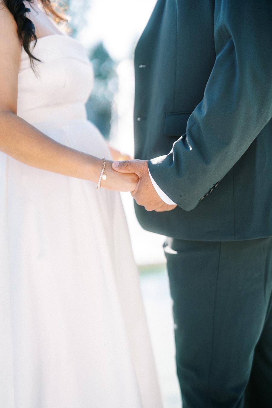 A couple holding hands in wedding attire, featuring a close-up of the bride's white dress and the groom's suit, symbolizing love and commitment on their wedding day.