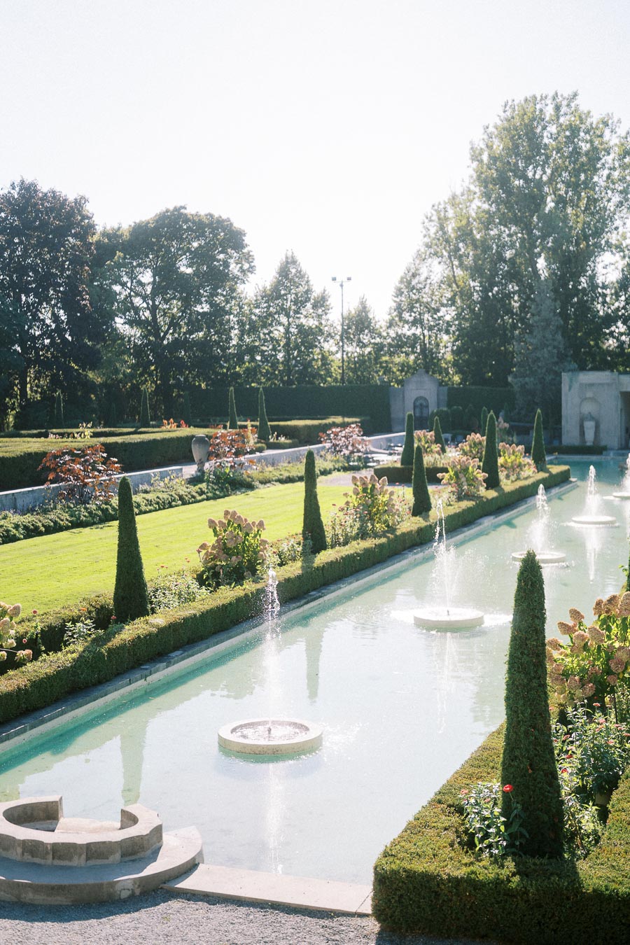 Luxurious formal garden with a long rectangular reflecting pool and multiple fountains surrounded by meticulously trimmed hedges, vibrant flowerbeds, and tall, symmetrical topiaries, under a clear blue sky.