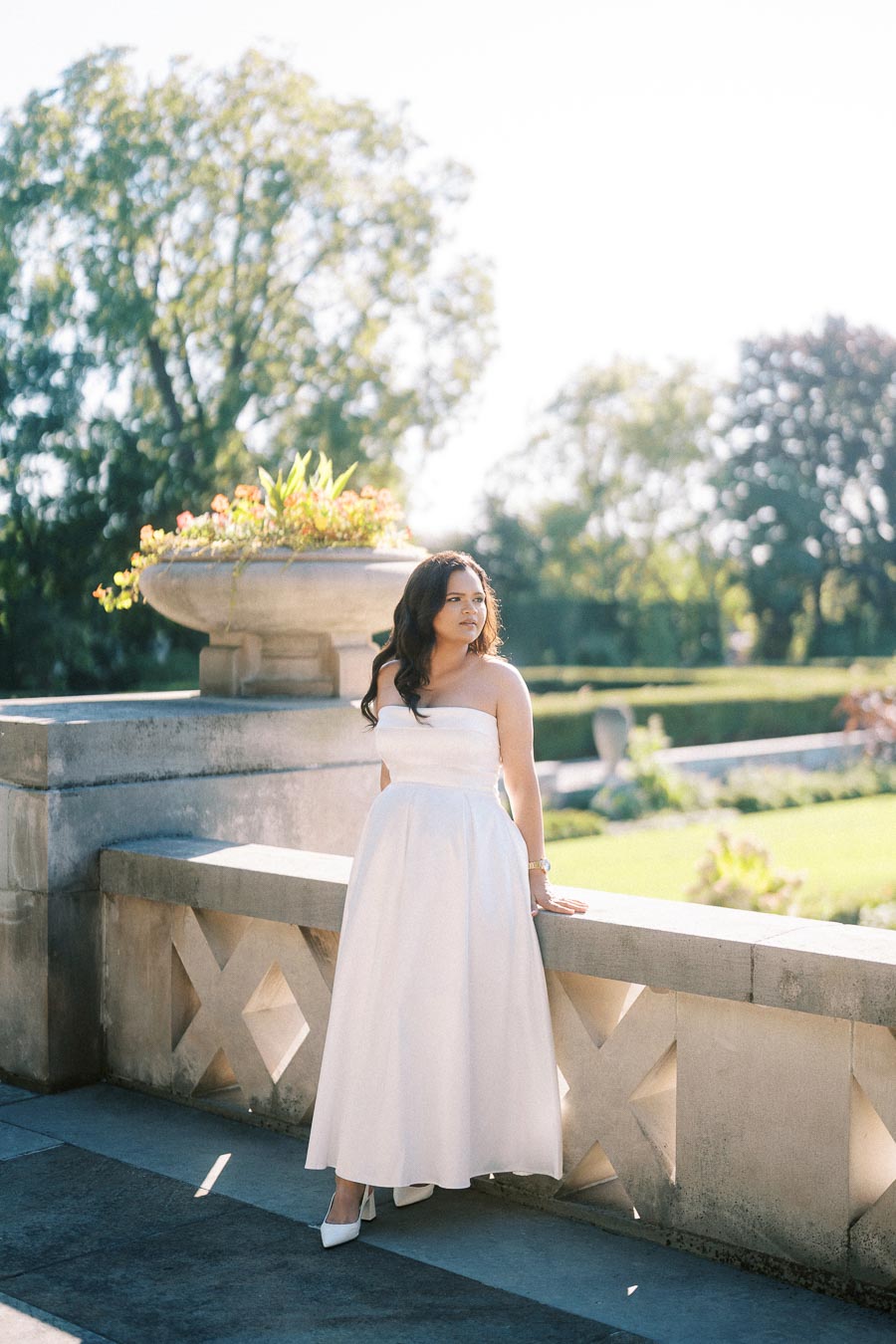 Woman in a white dress standing near a stone railing in a lush garden setting on a sunny day.