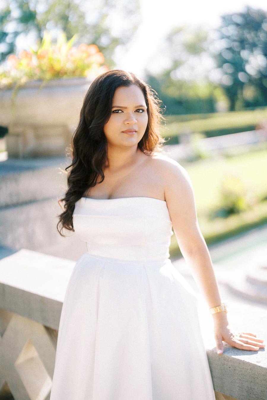 A woman in a white strapless dress stands by a stone railing in a sunny garden.