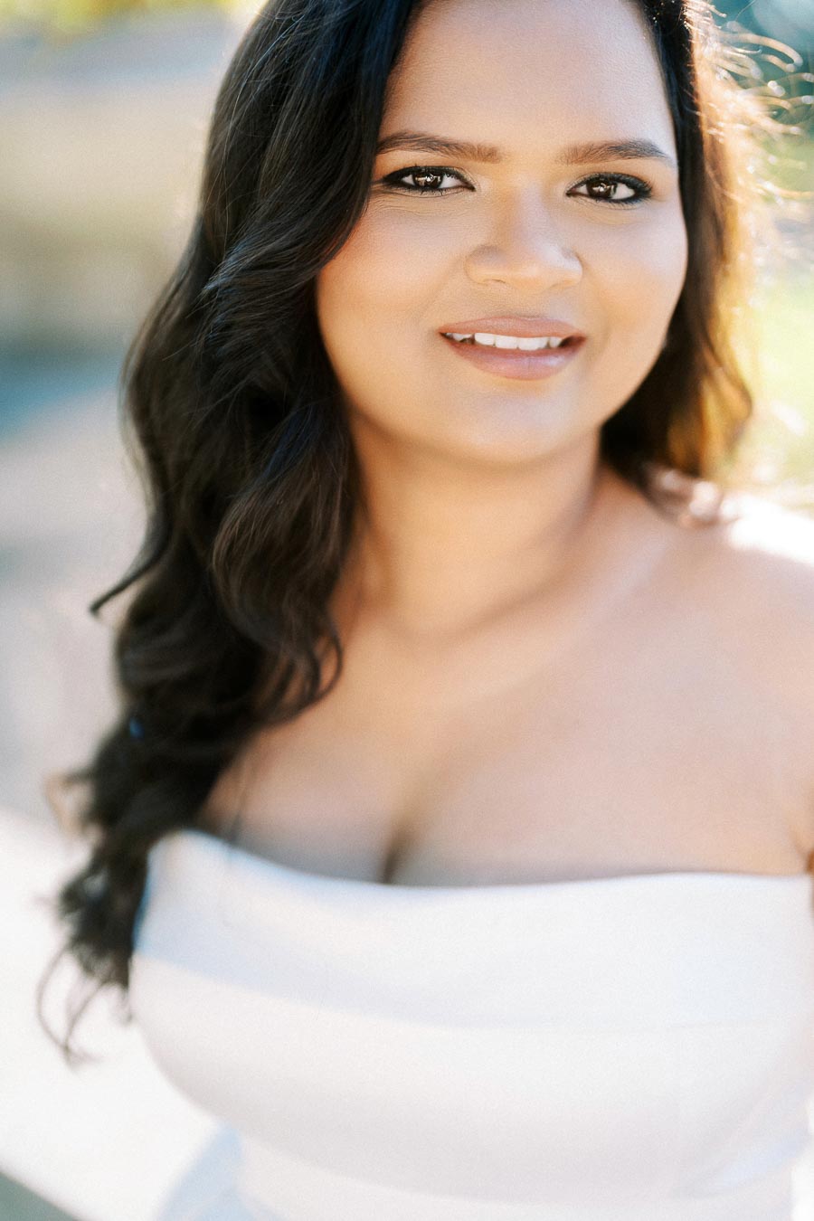 A woman with long dark hair smiles while wearing a white strapless dress outdoors.