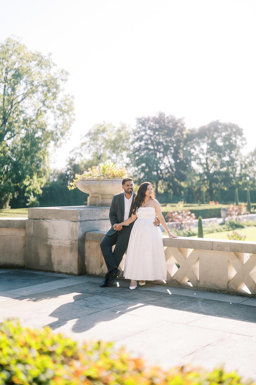 A couple in wedding attire enjoying a sunny day in a beautiful garden setting, with lush greenery and a decorative stone railing.