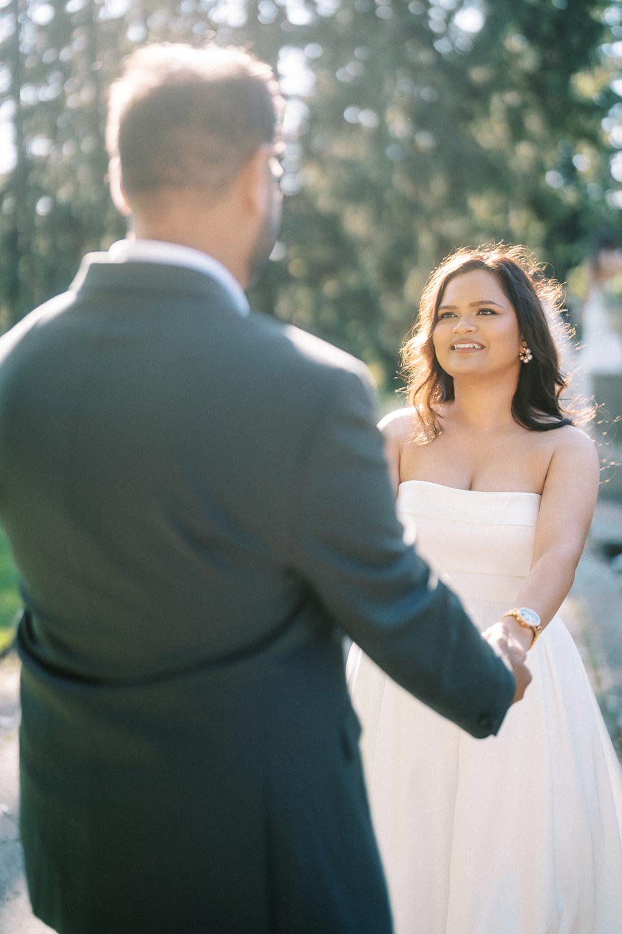 A couple holding hands outdoors, with the woman smiling in a white dress and the man wearing a suit, surrounded by a natural, sunlit background.