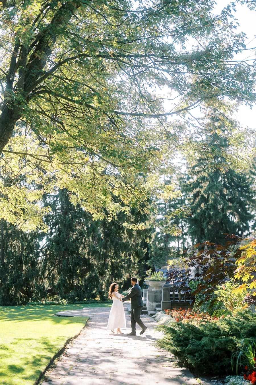 A couple joyfully dances on a sunlit garden path surrounded by lush greenery and blooming flowers, with tall trees providing shade on a bright day.