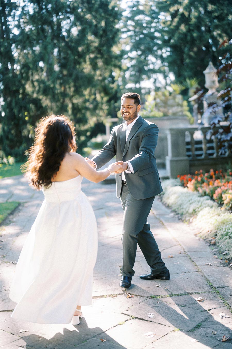 Happy couple dancing outdoors in a garden setting, with the woman in a white dress and the man in a suit, surrounded by lush greenery and flowers.