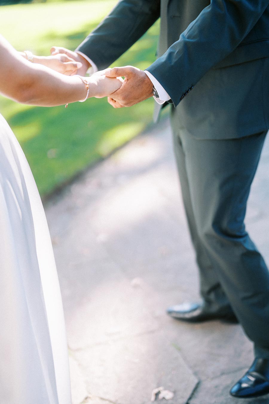 A couple holds hands outdoors, one dressed in a wedding gown and the other in a formal suit, symbolizing love and commitment in a sunlit garden setting.