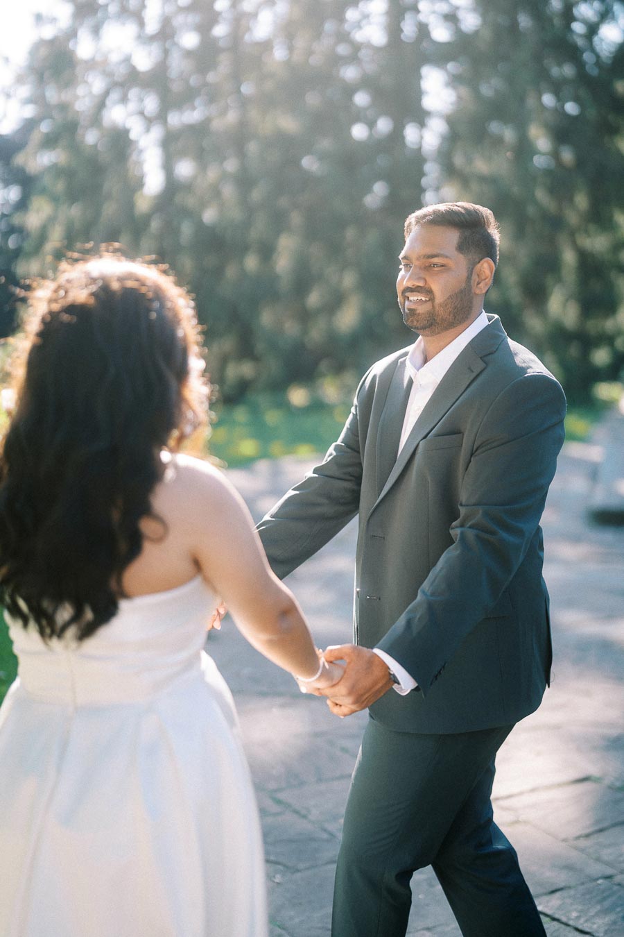 Couple holding hands and smiling outdoors on a sunny day, wearing formal attire; man in suit and woman in white dress, surrounded by lush greenery.