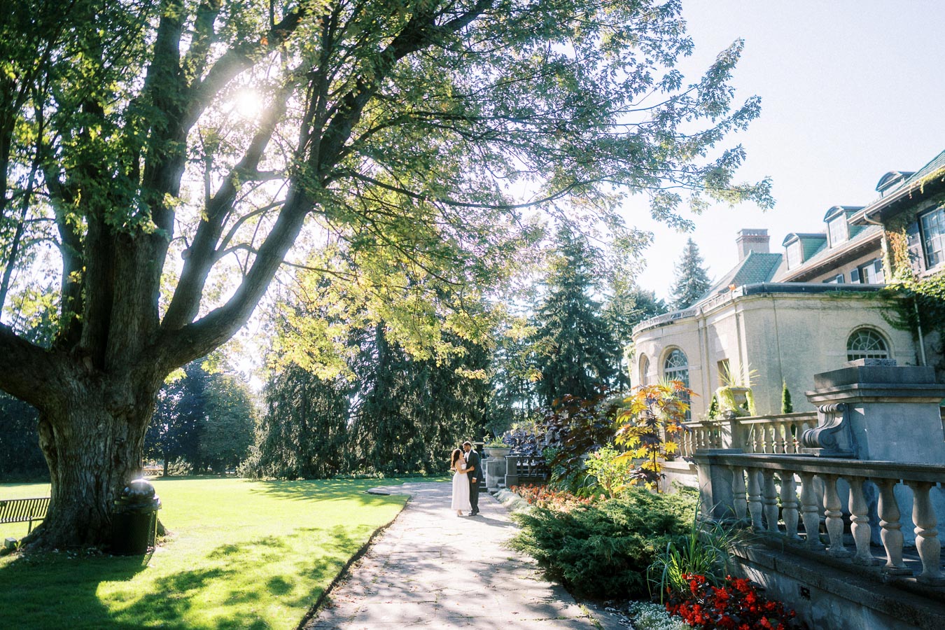 A couple embracing in a sunlit garden with a historic mansion in the background, surrounded by lush greenery and vibrant flowers.