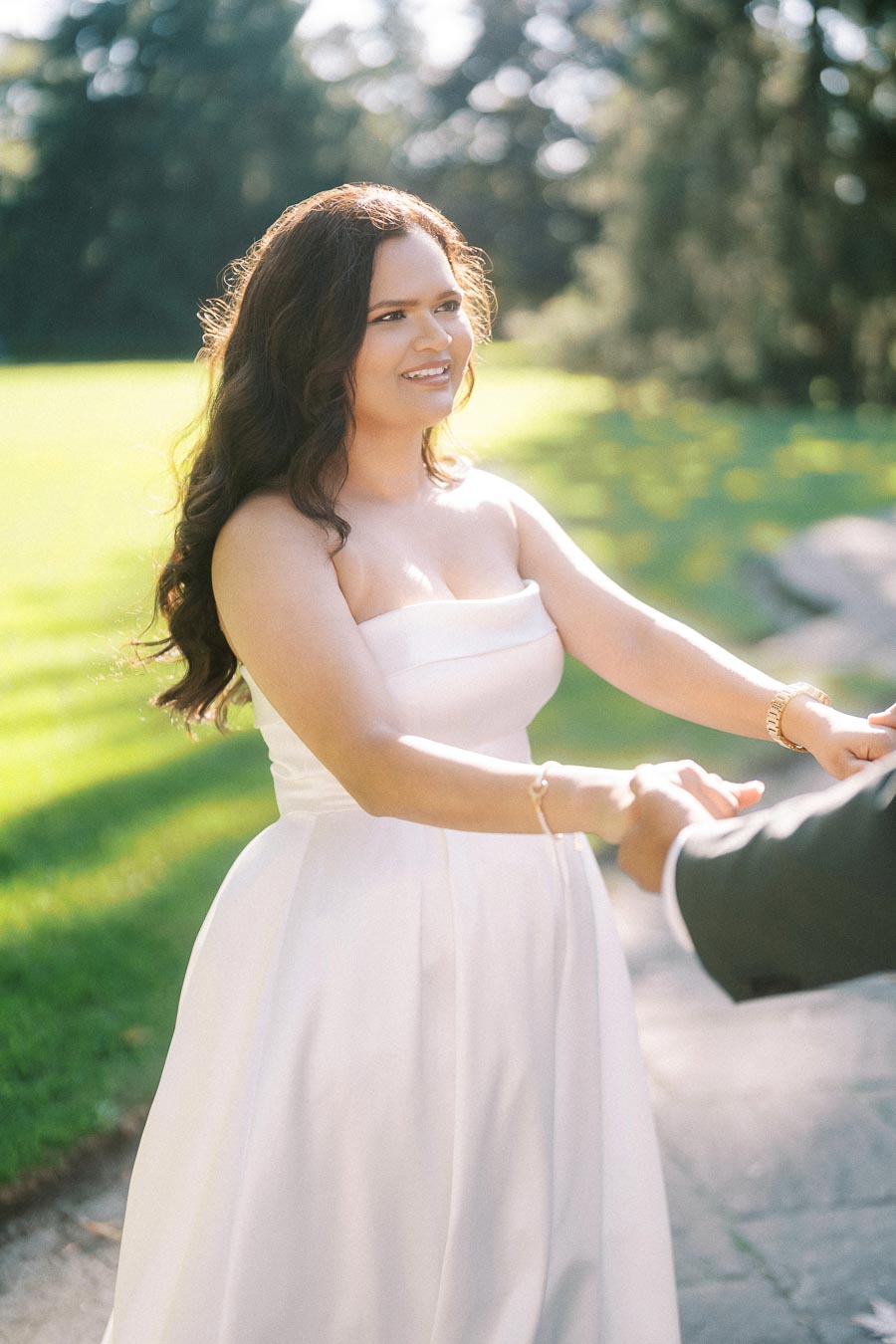 A bride in a white gown holds hands with a partner, smiling joyfully in a sunlit garden setting.