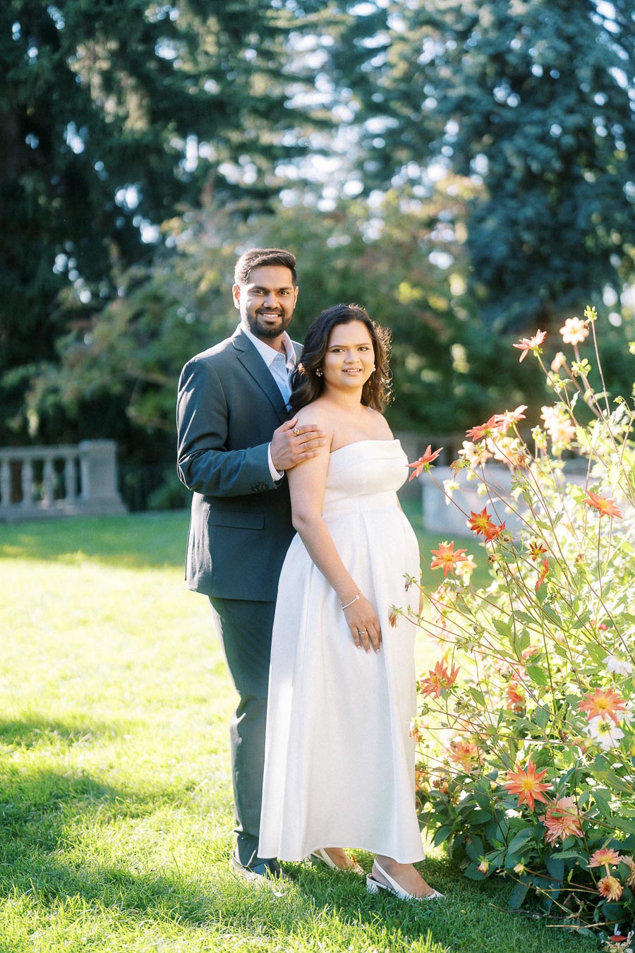 A couple posing together in a garden, with vibrant flowers in the foreground and lush trees in the background, capturing a romantic moment in elegant attire.
