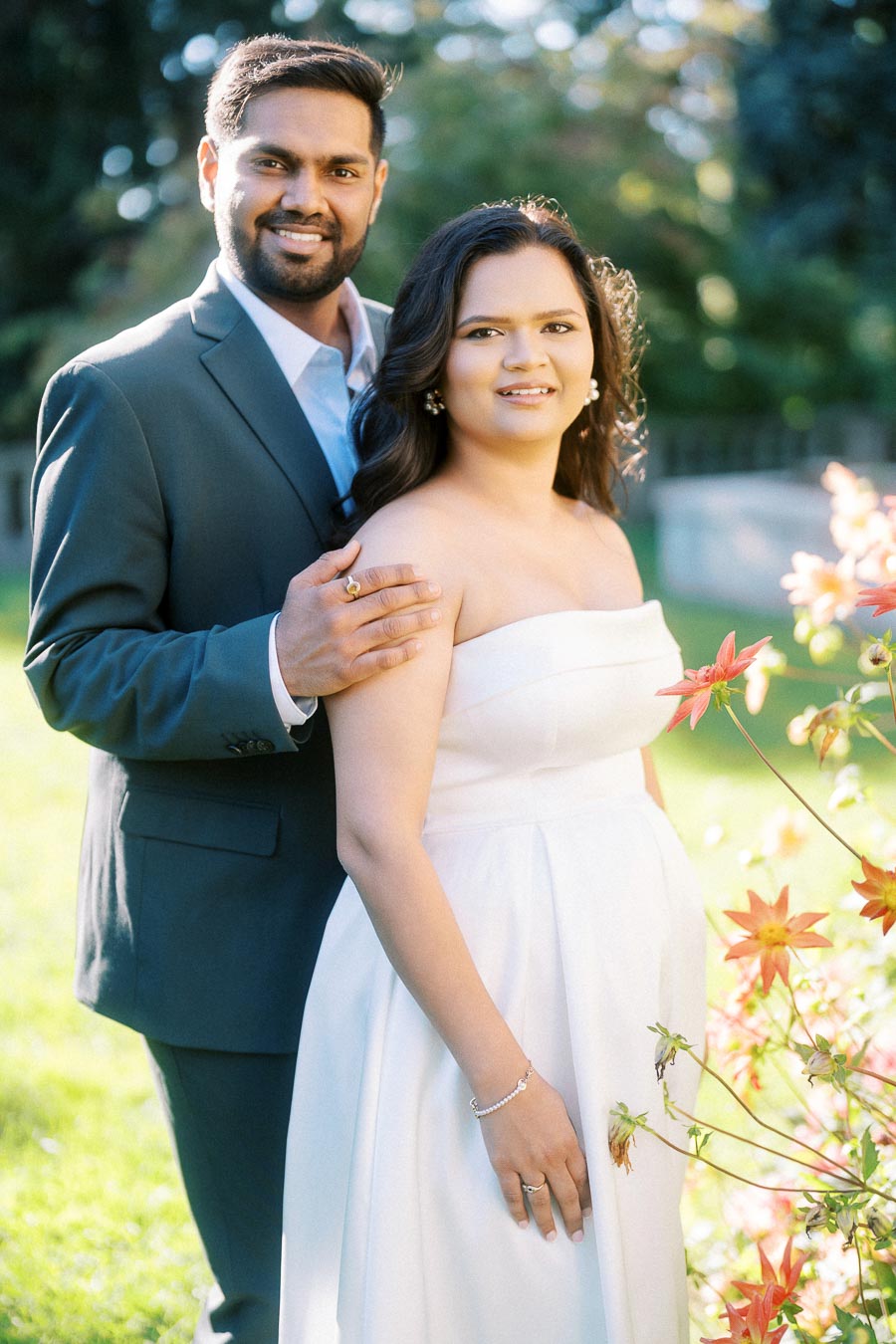 A couple in formal attire posing together outdoors, with the man wearing a dark suit and the woman in a white strapless dress, surrounded by flowers and greenery, smiling at the camera.