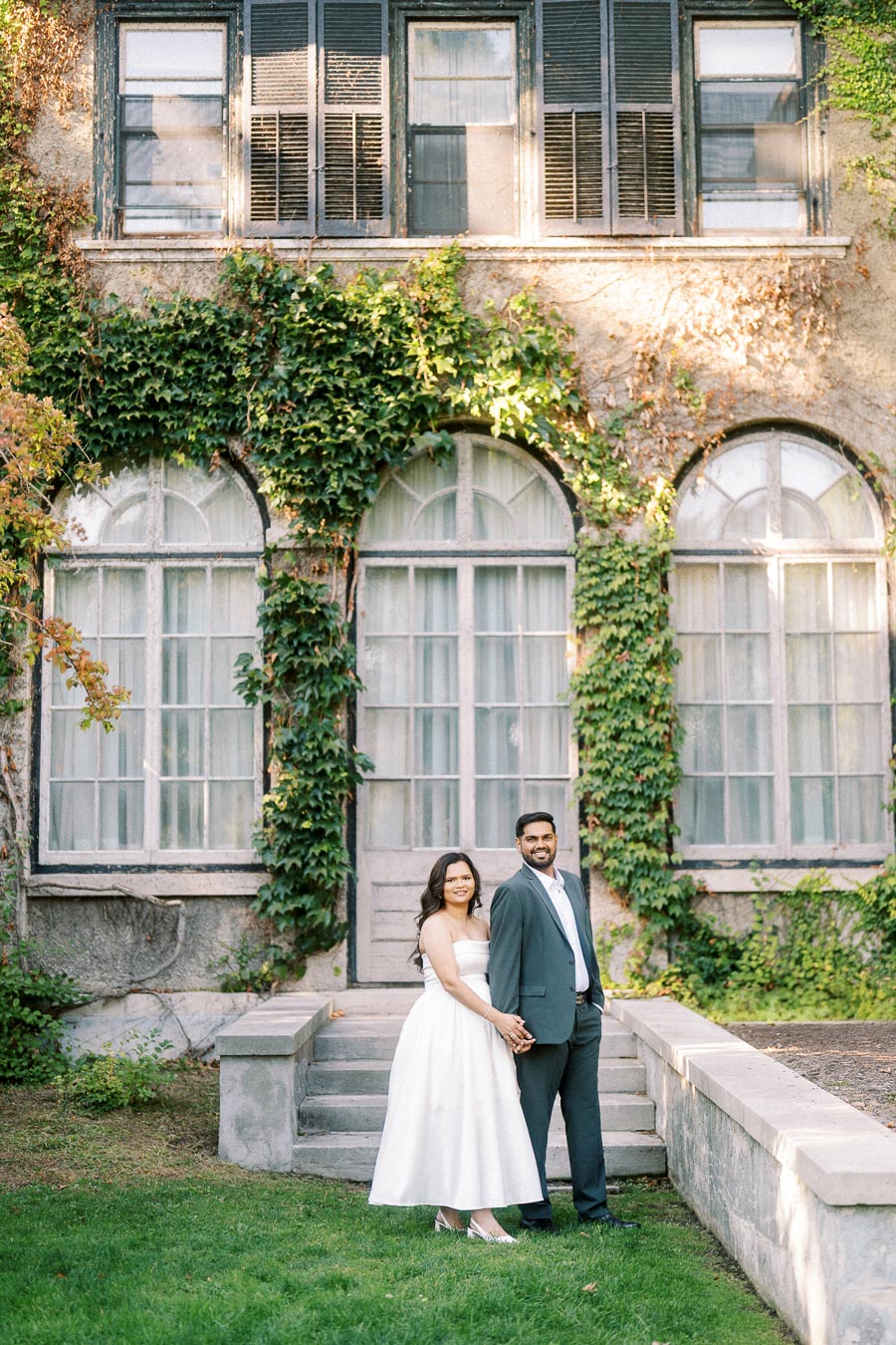 A couple standing in front of a historic building covered with ivy, posing for a wedding photo. The bride is wearing a white dress and the groom is in a dark suit, both smiling and holding hands in a picturesque garden setting.
