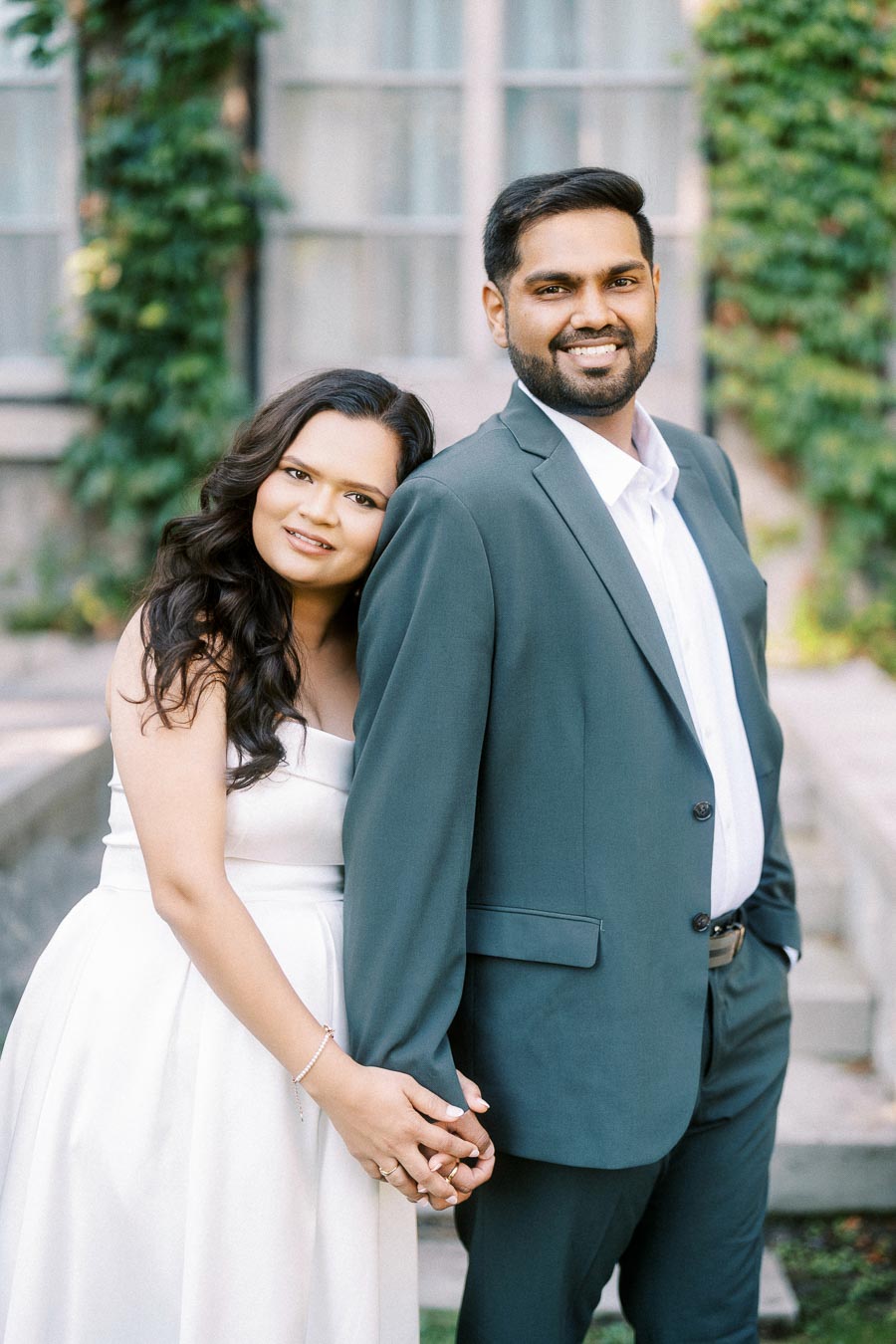 A couple poses outdoors, holding hands and smiling; the woman in a white dress leans against the man in a suit, with greenery and a windowed building in the background.