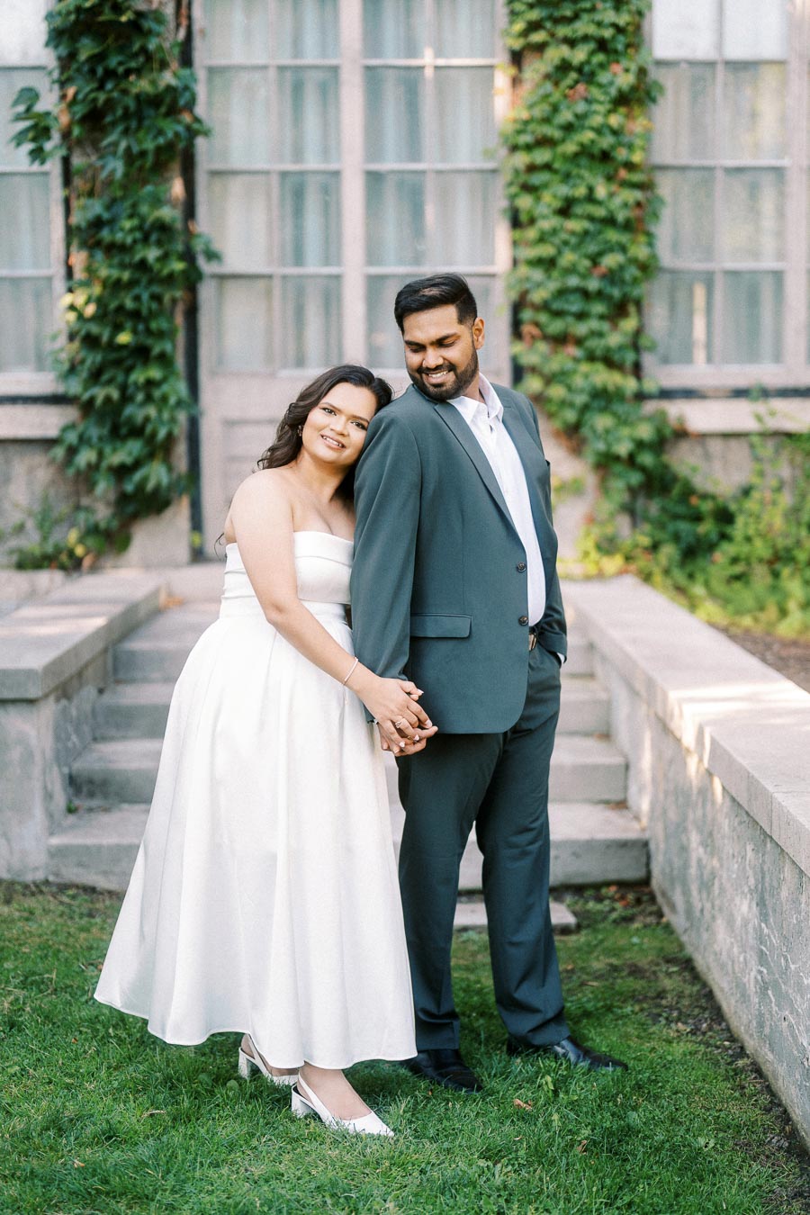 A couple in formal attire poses affectionately in a garden setting with ivy-covered walls and stone steps, exuding joy and elegance.