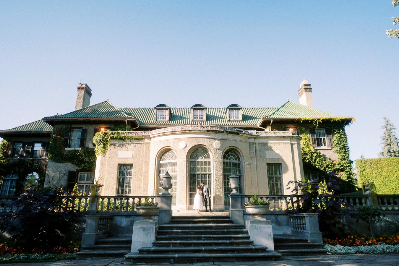 Elegant historic mansion with ivy-covered walls and a green roof, featuring a grand staircase and arched windows under a clear blue sky.
