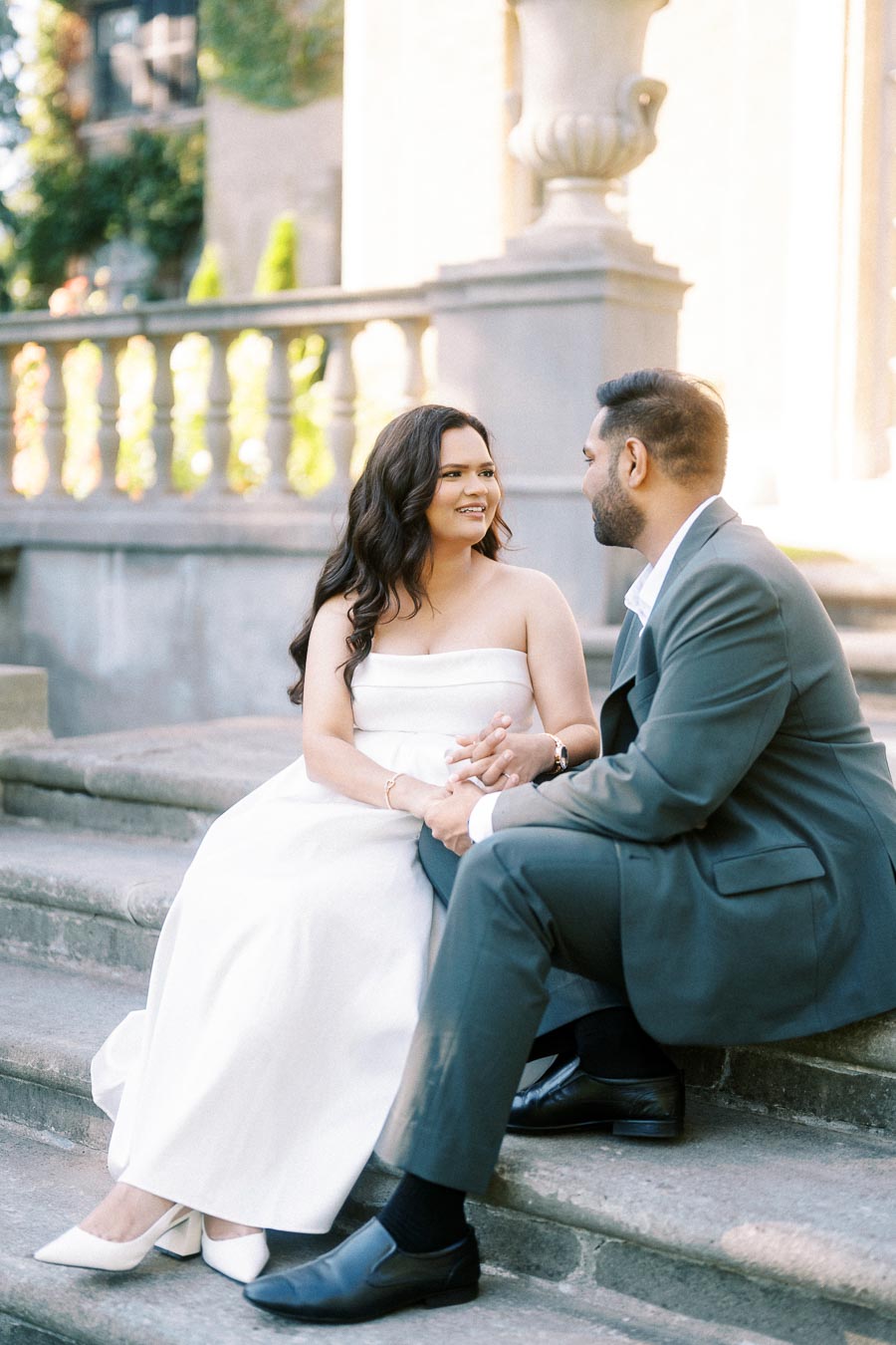 A couple in formal attire sitting on stone steps, holding hands and smiling at each other in an elegant outdoor setting.