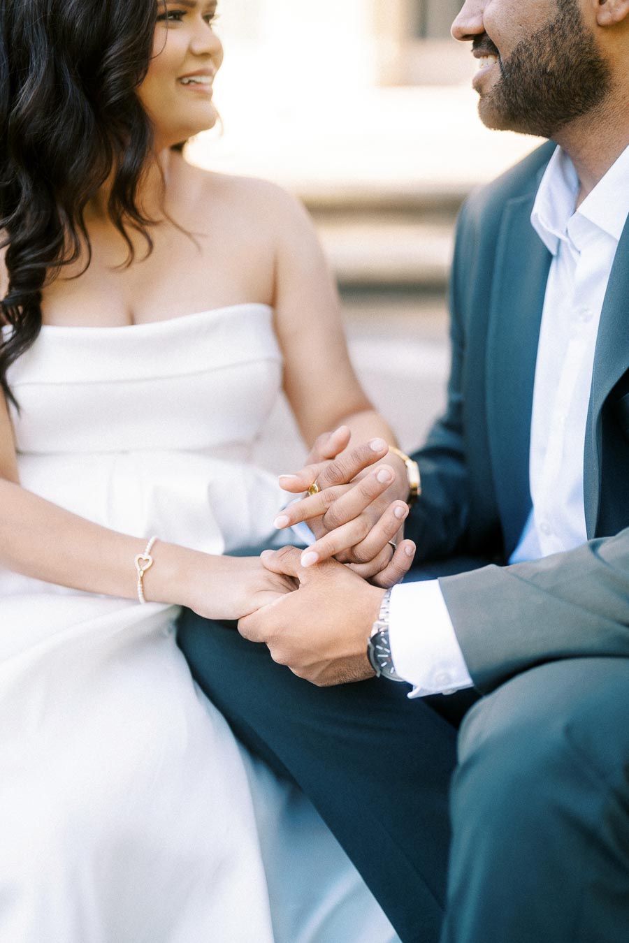 A couple in elegant attire holding hands, with the woman wearing a white dress and a gold heart bracelet, expressing love and connection.