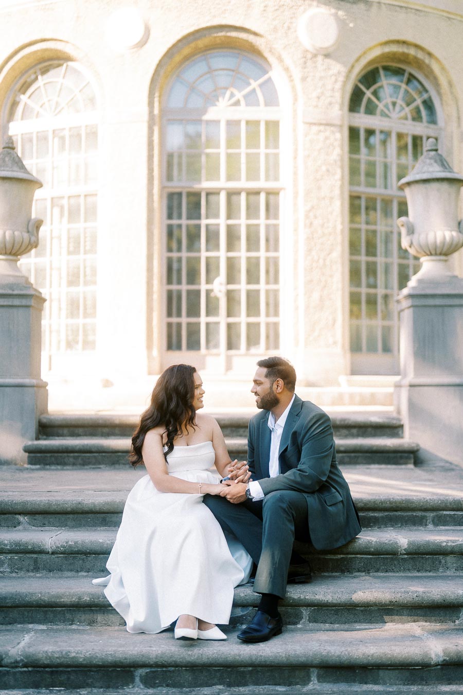 A couple sitting on stone steps in front of an elegant building with arched windows, dressed in formal attire, the woman in a white gown and the man in a dark suit, holding hands and smiling at each other.