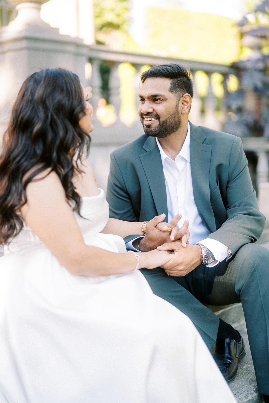 A couple sitting on steps, holding hands and smiling at each other, dressed in formal attire.