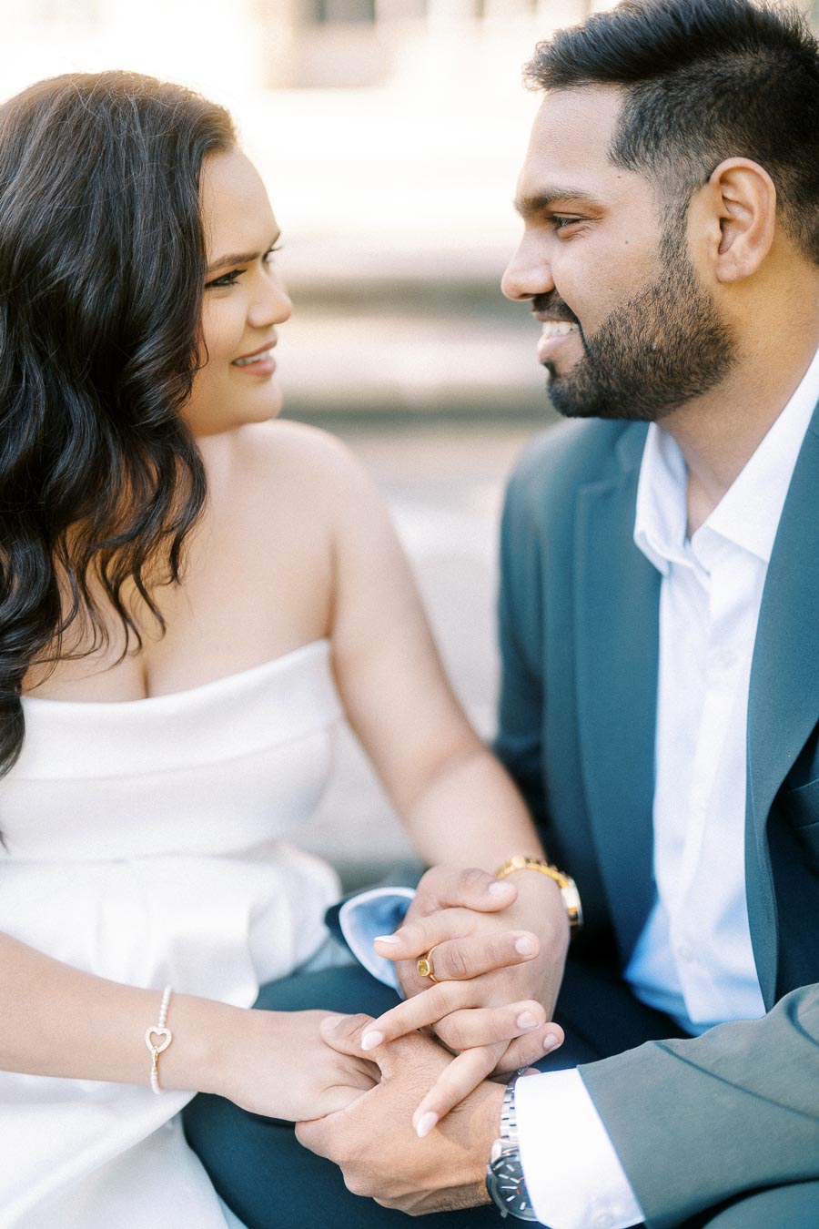 A couple in formal attire holding hands and smiling lovingly at each other, with the woman in a white dress and the man in a suit, captured in a soft-focus outdoor setting.