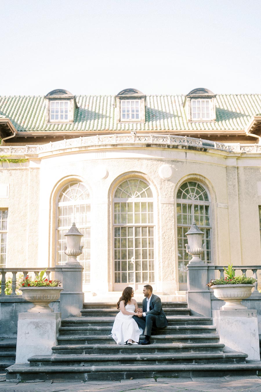 A couple in elegant attire sitting on stone steps in front of a historic building with large arched windows and green tiled roof, surrounded by decorative urns with plants.