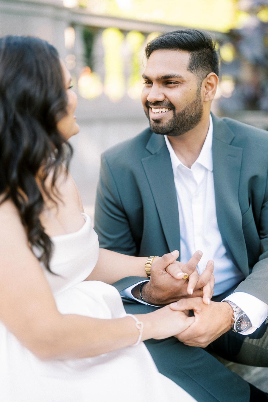 Engaged couple holding hands and smiling at each other, with the man in a green suit and woman in a white dress, capturing a joyful moment during their photoshoot in an outdoor setting.