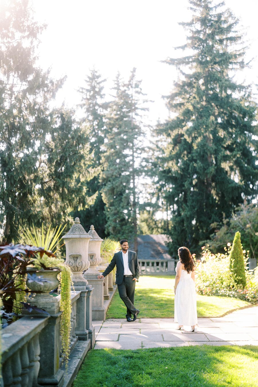 A couple enjoying a romantic moment in a sunny garden with tall evergreen trees and stone balustrades, creating a serene outdoor setting.