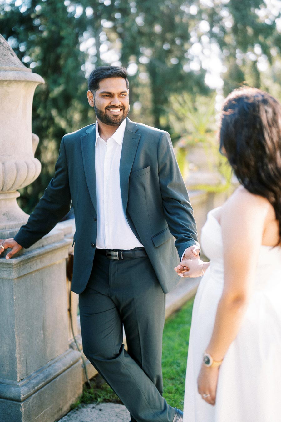 A couple holding hands in an outdoor setting, with the man in a dark suit smiling at the woman in a white dress, surrounded by greenery and stone architecture.