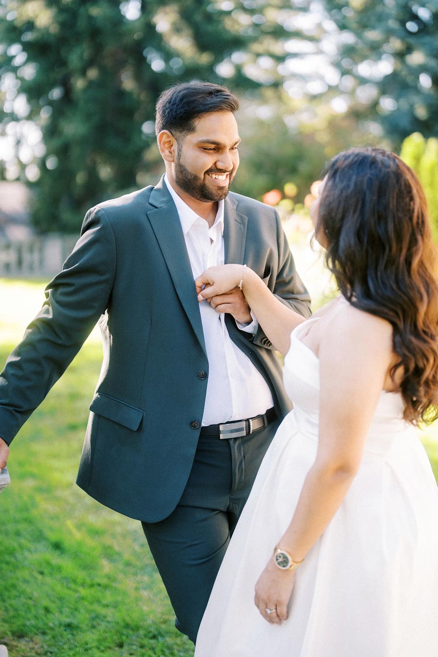 A couple dressed in formal attire sharing a joyful moment outdoors, with the man in a dark suit and the woman in a white dress, set against a backdrop of lush greenery and sunlight.