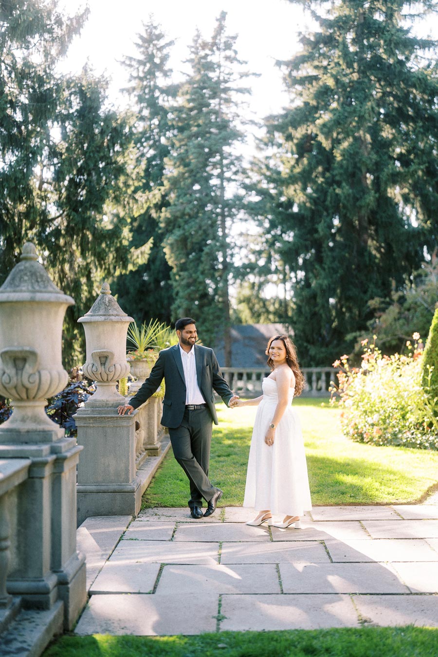 A couple standing in a beautiful garden setting, with the man in a suit and the woman in a white dress, surrounded by lush greenery and ornate stone railings.